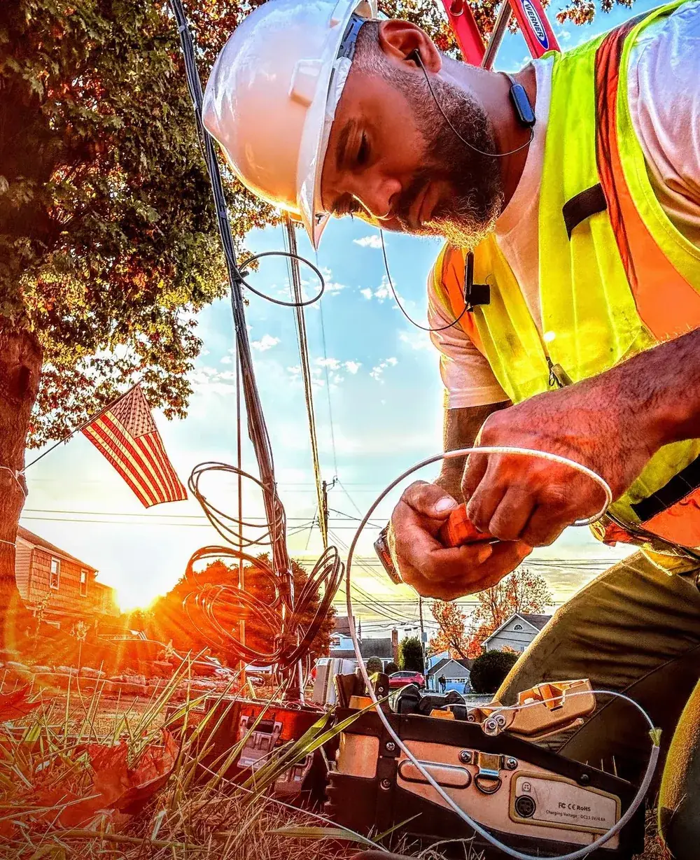 APH Fiber technician in a hard hat and safety vest splicing fiber at sunset